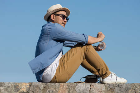 Asian tourist man open a bottle of water  while sitting against blue sky.の写真素材