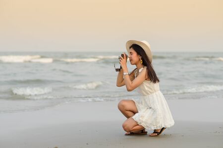 Asian woman wearing hat with white dress taking photo on the beach.の写真素材