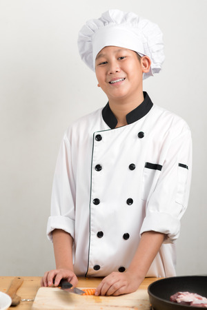 Asian preteen boy in chef uniform chopping carrot on table.の写真素材