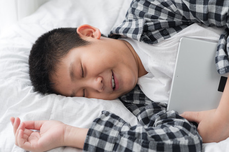 Asian preteen boy holding tablet in his arm while sleeping on bed at home.の写真素材