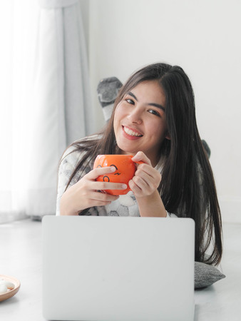 Happy Asian girl lying on floor and drinking hot chocolate at home,lifestyle in winter concept.