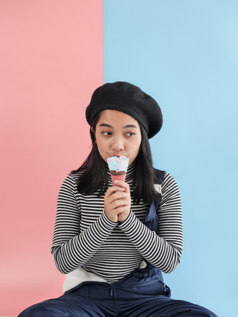 Happy Asian woman eating ice cream cone with blue and pink background.