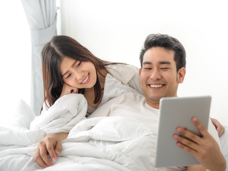 Happy Asian couple using laptop and tablet together on bed at home, lifestyle concept.の写真素材