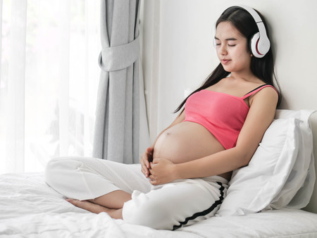 Beautiful Asian pregnant woman using headphone on her belly at home, lifestyle concept.の写真素材