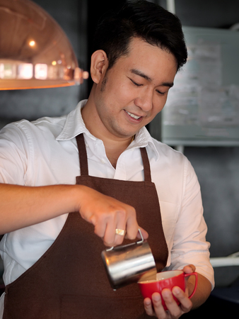 Happy Asian barista pouring milk into red coffee cup for latte art.の写真素材