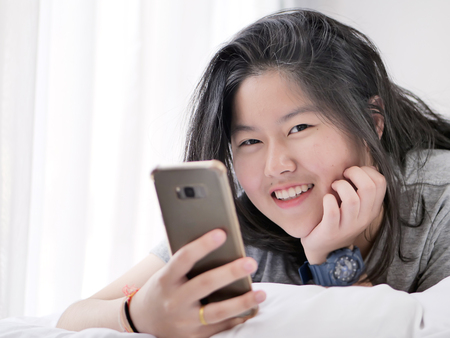 Happy Asian teenager girl using smartphone on bed at home, lifestyle concept.の写真素材
