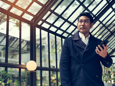 Happy Asian businessman in blue coat using smart phone in cafe, lifestyle concept.の写真素材