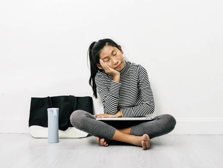 Sleeping Asian girl using laptop while sitting on floor with fabric handbag and water bottle,...