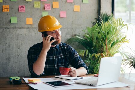 Engineer talking with phone and drinking coffee in office, colorful sticky notes on cement wall background.の写真素材