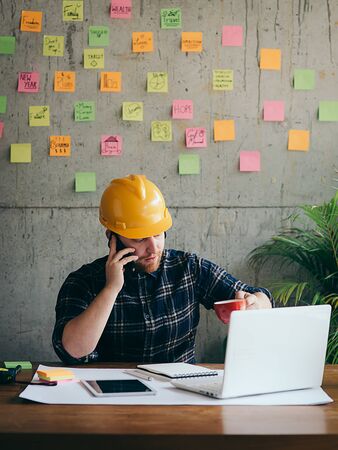 Engineer talking with phone and looking at laptop in office, colorful sticky notes on cement wall background.の写真素材