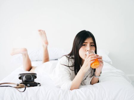 Asian woman drinking a glass of orange juice on bed at home, lifestyle concept.