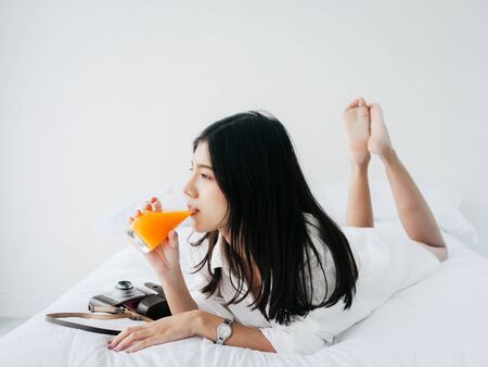 Asian woman drinking a glass of orange juice on bed at home, lifestyle concept.の写真素材