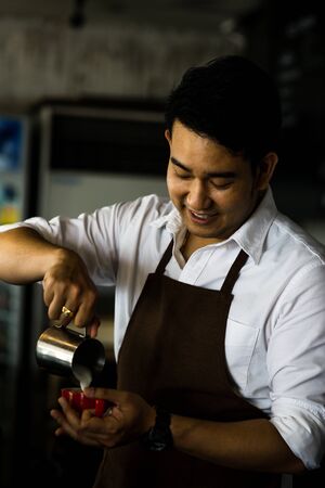 Happy Asian barista pouring milk into red coffee cup for latte art.の写真素材