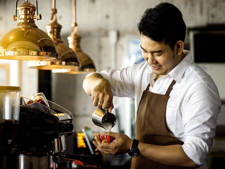 Happy Asian barista pouring milk into red coffee cup for latte art.の写真素材