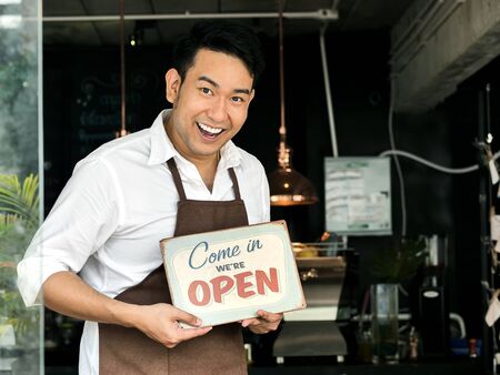 Cheerful Asian barista man holding welcome board in front of coffee shop.