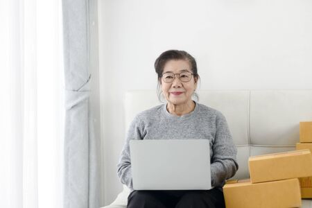 Asian senior woman using laptop while selling online with packaging, work at home concept.