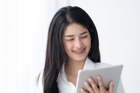 Beautiful Asian woman in white shirt using tablet near window in the morning.