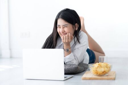 Asian woman in white shirt laying on floor and using laptop at home.