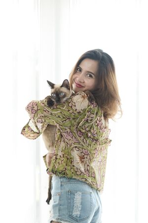 Smiling Asian woman holding her cat with window light at home.の写真素材