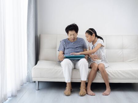 Asian senior woman and girl using tablet  and eating orange at home, lifestyle concept.の写真素材