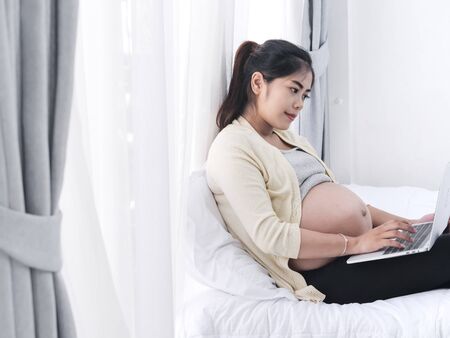 Asian pregnant woman using laptop on bed at home, lifestyle concept.の写真素材