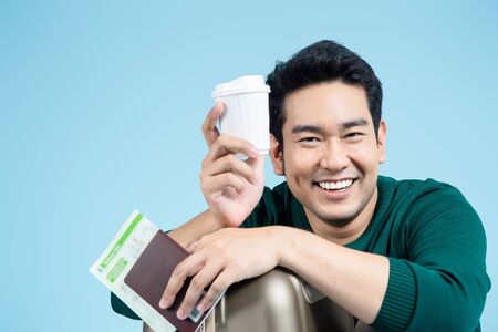 Happy Asian tourist sitting and holding  take away coffee, passport , map on blue background,