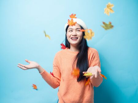 Happy Asian woman wearing sweater and playing with dropping maple leafs on blue background, motion...