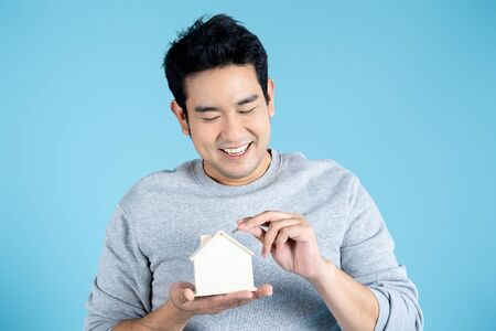 Happy Asian man with wooden house piggybank on blue background.の写真素材