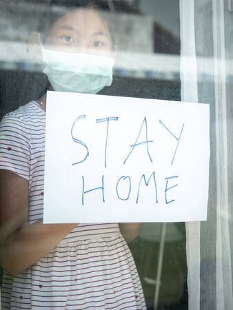 Asisan girl wearing mask holding paper message STAY HOME, during COVID-19.の写真素材