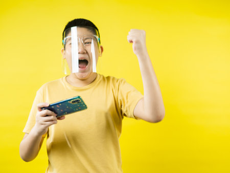 Asian preteen boy wearing face shield and playing game by smartphone with yellow background, new normal concept.の写真素材