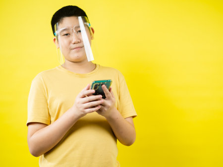 Asian preteen boy wearing face shield and playing game by smartphone with yellow background, new normal concept.の写真素材
