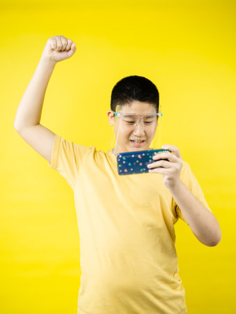 Asian preteen boy wearing face shield and playing game by smartphone with yellow background, new normal concept.の写真素材