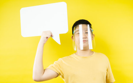 Asian preteen boy wearing face shield holding blank speech bubble on yellow background.の写真素材