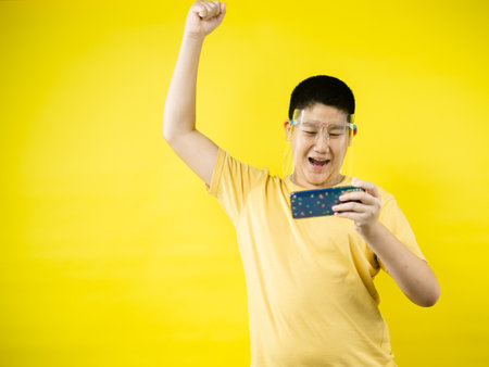Asian preteen boy wearing face shield and playing game by smartphone with yellow background, new normal concept.の写真素材