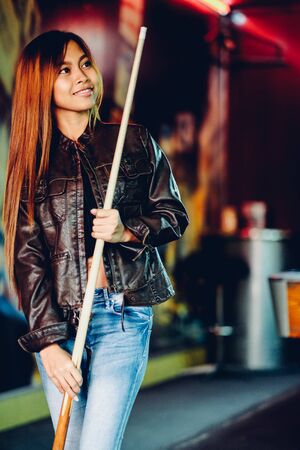Young beautiful girl wearing leather jacket in a billiard club, with cue stick preparing for the gameの写真素材