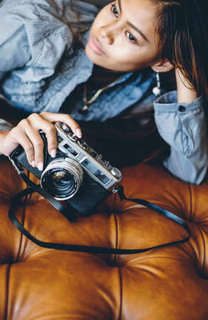 Gorgeous girl lying on leather sofa with photo camera in her handsの写真素材