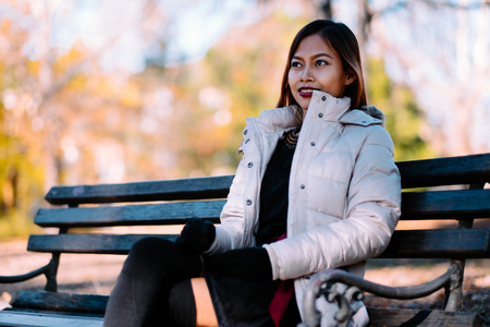 Portrait of young beautiful woman sitting on the bench in stylish warm outfit in sunny autumn day in park. Casual lifestyle in the cityの写真素材