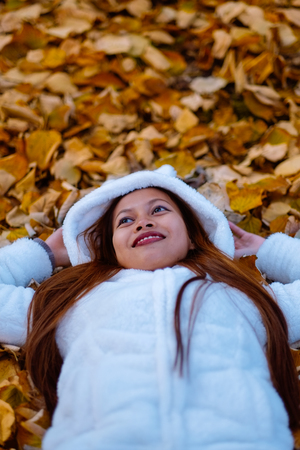 Autumn girl playing in city park. Portrait of an autumn woman lying over leaves and smiling outside in fall forest. Beautiful energetic Asian young womanの写真素材