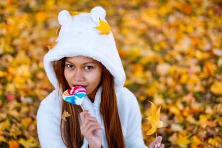Young woman in park on sunny autumn day, smiling, holding leaves and candy. Cheerful beautiful girl in white sweaterの写真素材