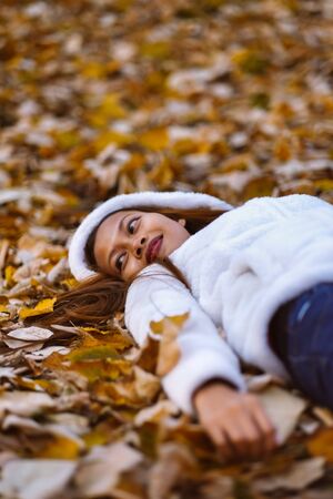 Autumn girl playing in city park. Portrait of an autumn woman lying over leaves and smiling outside in fall forest. Beautiful energetic Asian young womanの写真素材