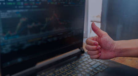 Hand show thumbs up sight with stock market data on a laptop screen at home