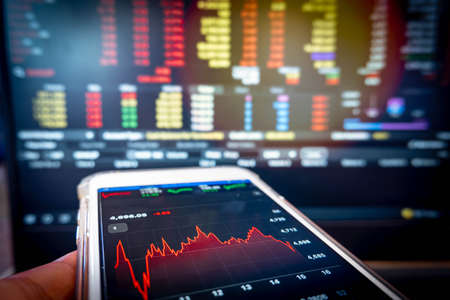 Businessman holding phone checking stock market data on computer screen background.