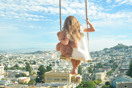 Barefoot girl in white dress hugging teddy bear, flying on vintage swings high above city, view from behindの写真素材