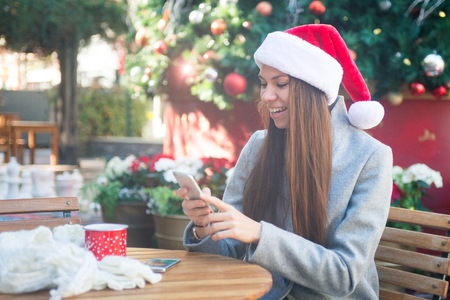 Smiling woman in Santa hat texting near Cristmas tree in cafe outsideの写真素材