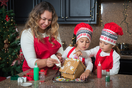 Happy Mom and twin girls in red making Christmas gingerbread houseの写真素材