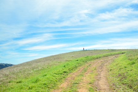 Trail in rural landscape with hiker on top of the hill, green grass hills under cloudy skyの写真素材