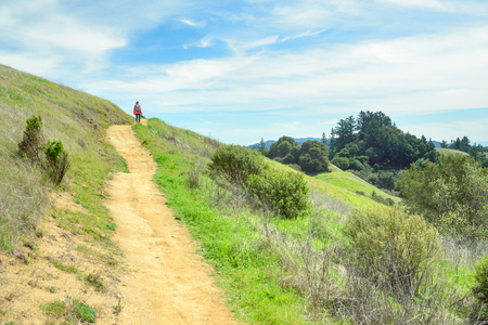 Hiker woman in red jacket with backpack on trail in beautiful landscape with green hills and cloudy sky, travel and backpackingの写真素材