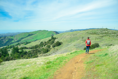Hiker woman in red jacket with backpack on trail in beautiful landscape with green hills and cloudy sky, travel and backpackingの写真素材