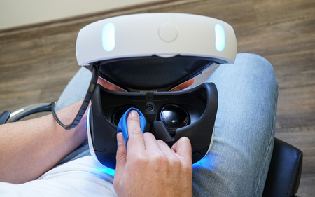 Man cleaning virtual reality glasses, sitting on the chair in the room, wearing white t-shirt and...