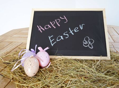 Chalkboard with text Happy Easter and decorative eggs on dry grass and a wooden background